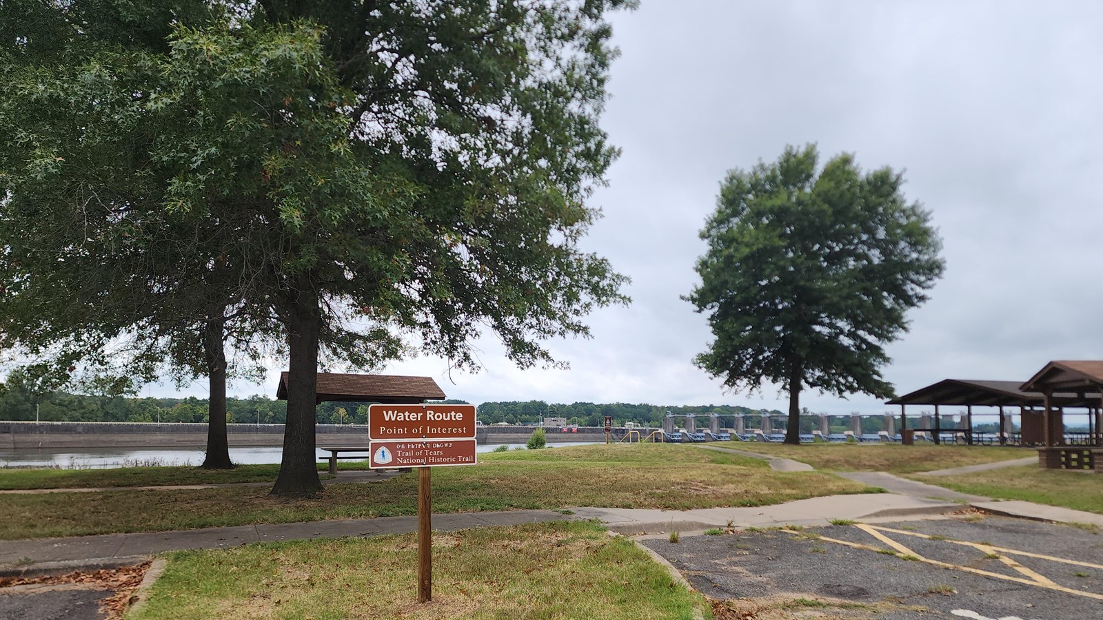 A brown sign in a grassy patch in a parking lot with distant trees in a park setting next to a river