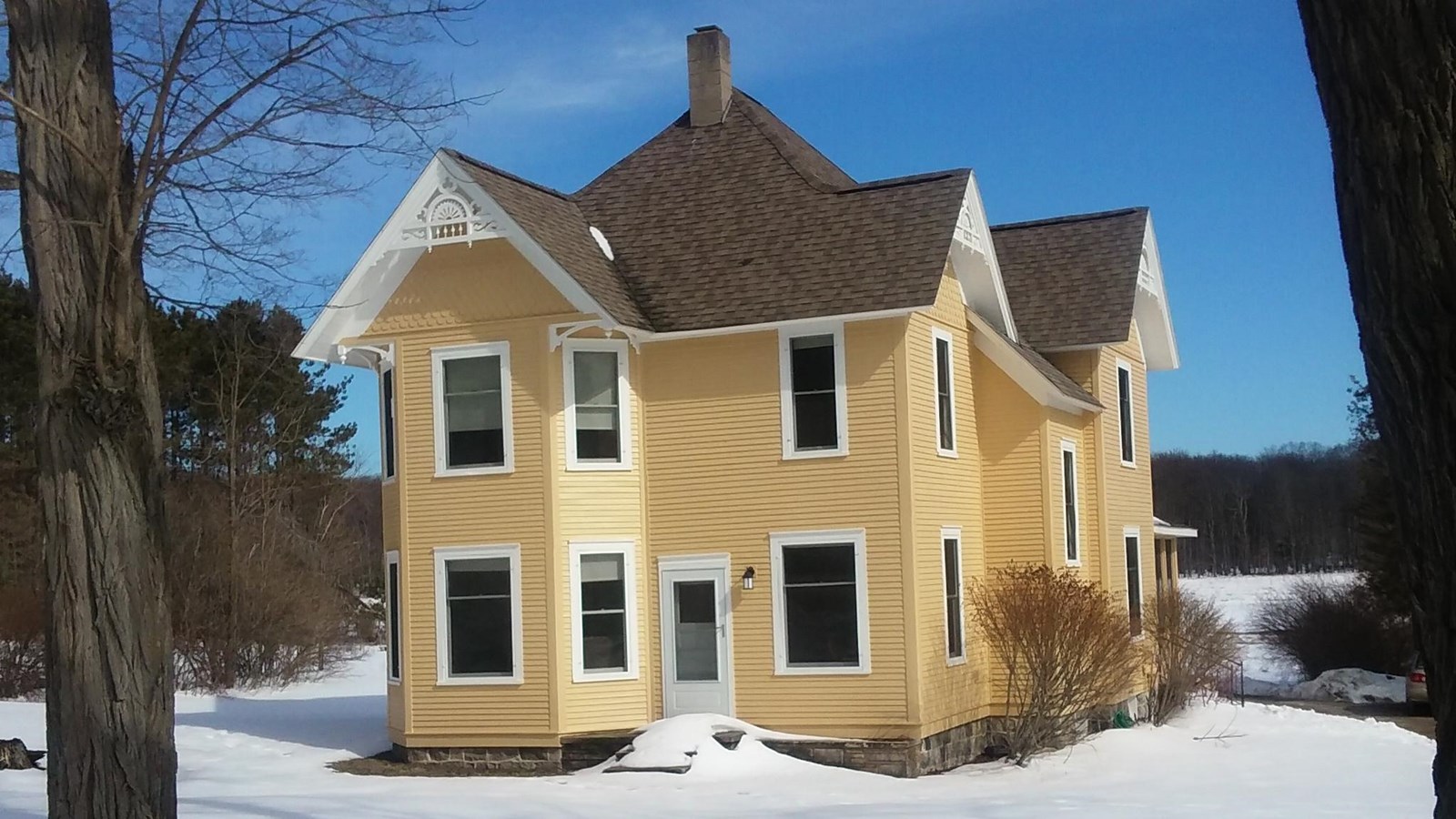 Two-story Victorian home in subtle yellow stands between two trees amongst a small blanket of snow. 