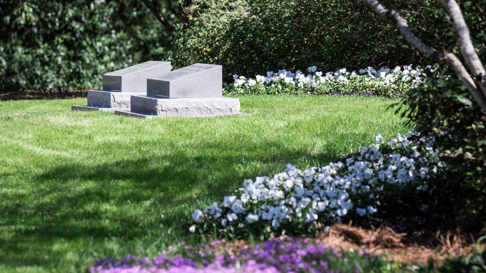 Two simple grey headstones side-by-side surrounded by a garden of trees and flowers. 