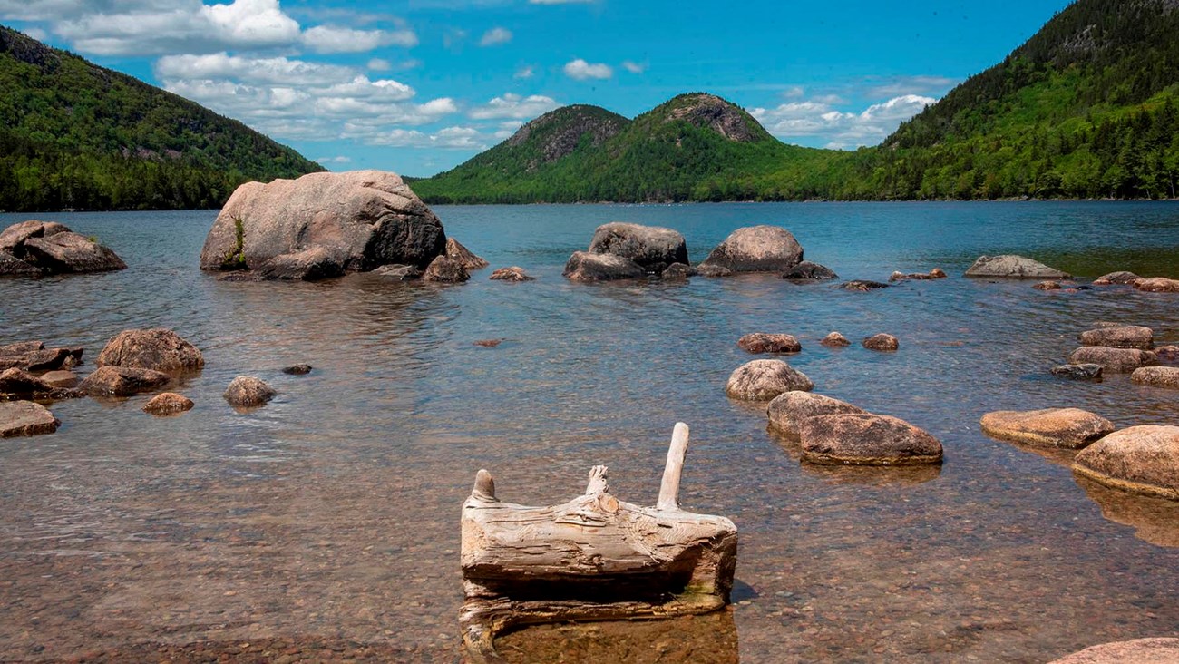 The placid surface Jordan Pond with exposed boulders and a log protruding from the water.