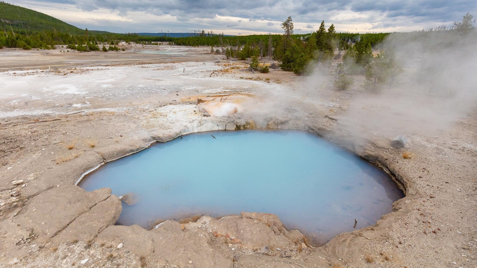 A steamy hot spring with milky blue waters sits on the barren landscape in a thermal basin.