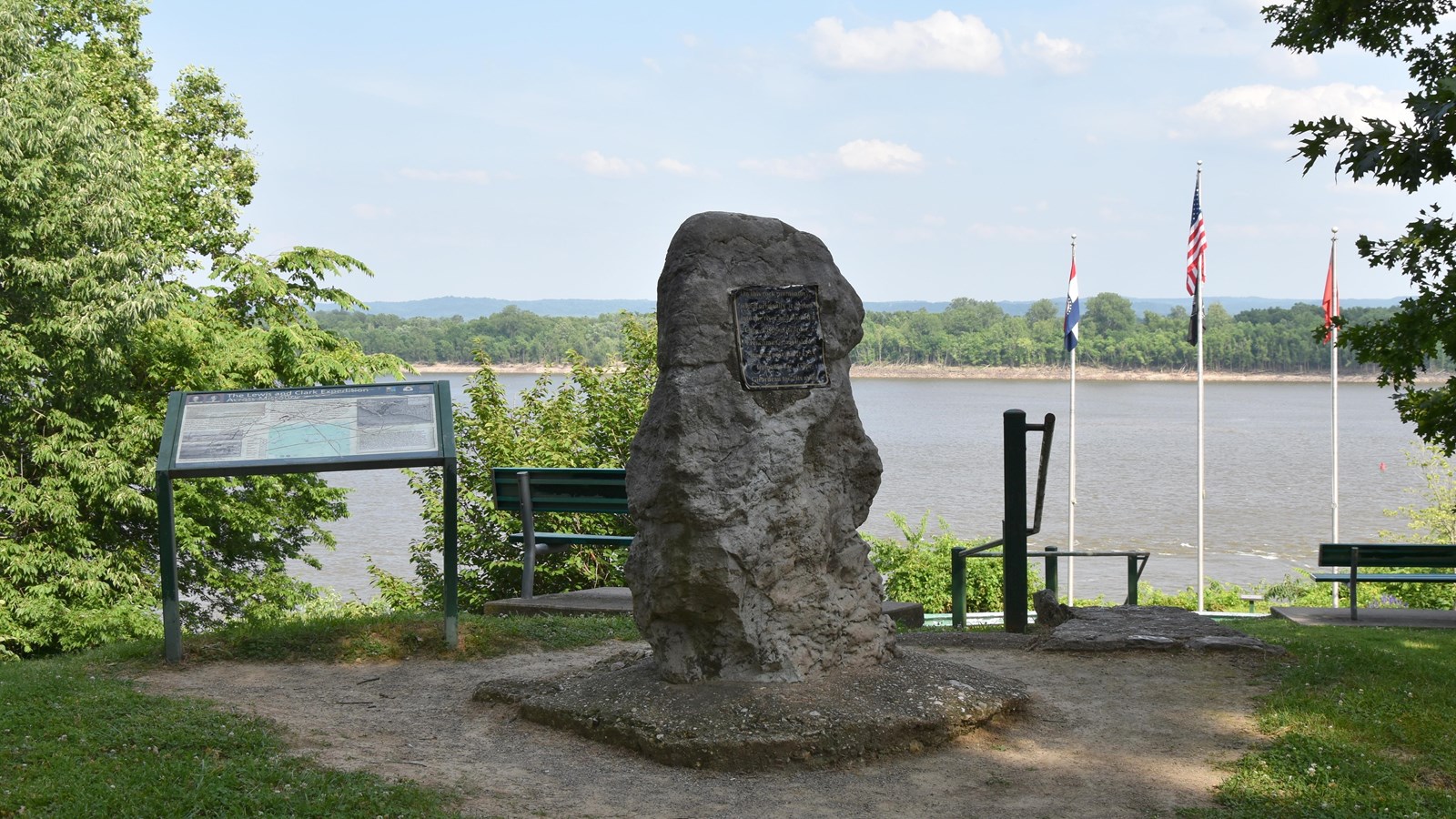 A stone monument with a mounted plaque stands at a shaded river overlook, framed by trees, benches