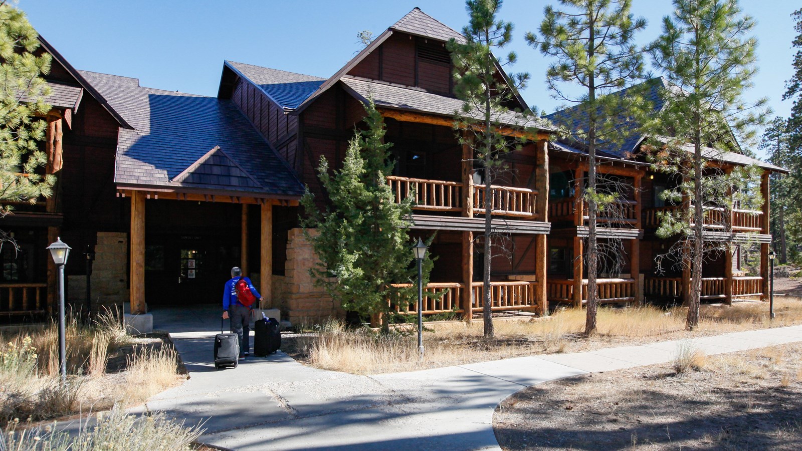 A man wheels his luggage into a large rustic style building with balconies and a metal roof.
