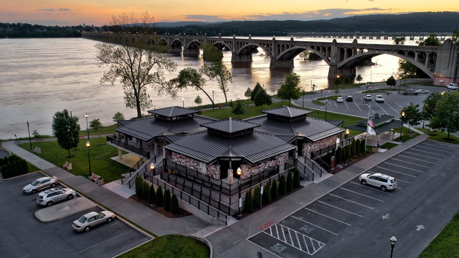 Aerial view of a building complex by a river with an arch bridge in the background at sunset.
