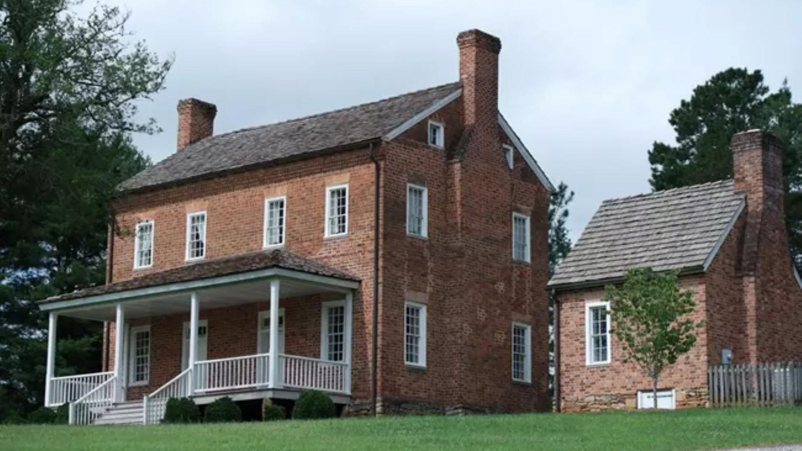 A two-story brick house with a small brick side house, trees and green grass surround the house. 