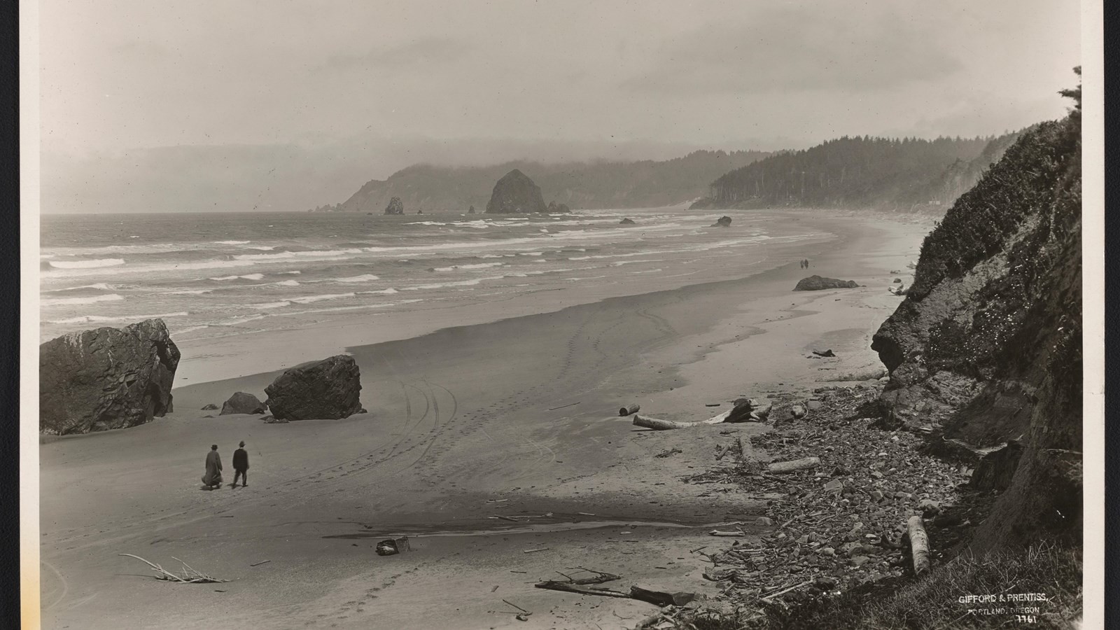 Sweeping landscape of sandy beach, ocean with white-capped waves, rocky outcroppings in the water.