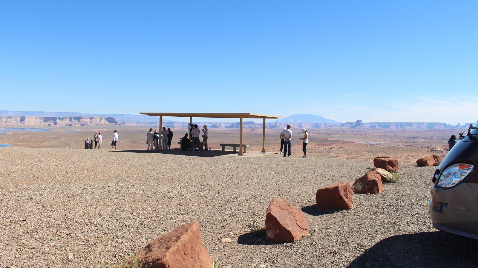 A group of people standing under a shade shelter near a gravel parking lot