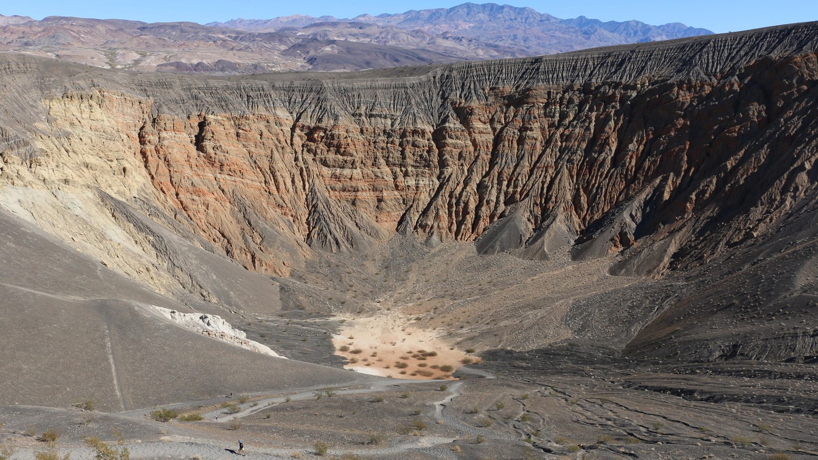 A visitor hikes in the bottom of a colorful crater with black cinders on the rim and eroded walls.