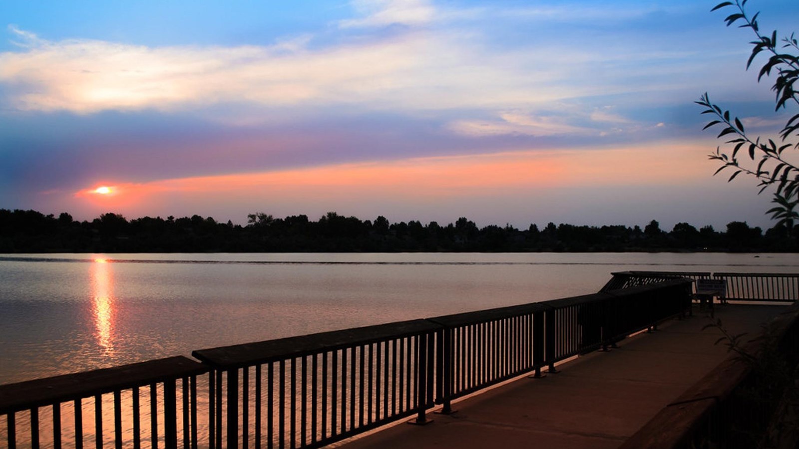 Sunset paints the sky above a tree-lined lake and boardwalk with pink and blue.