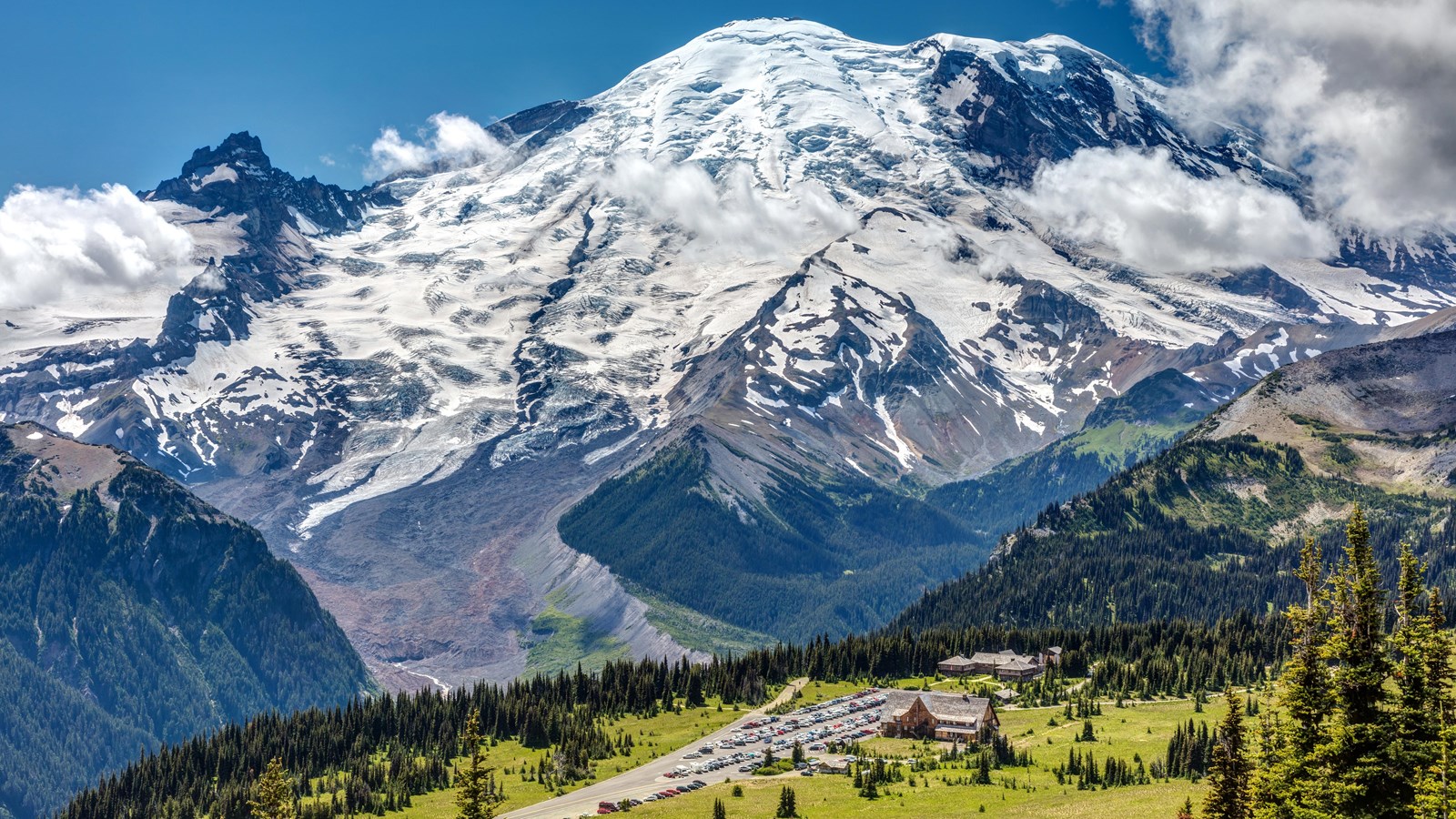Mount Rainier with meadows dotted with wildflowers and trees leads to a parking lot and buildings.