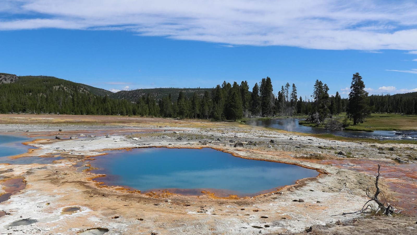 A turquoise blue hot spring sits in a thermal area near a river.