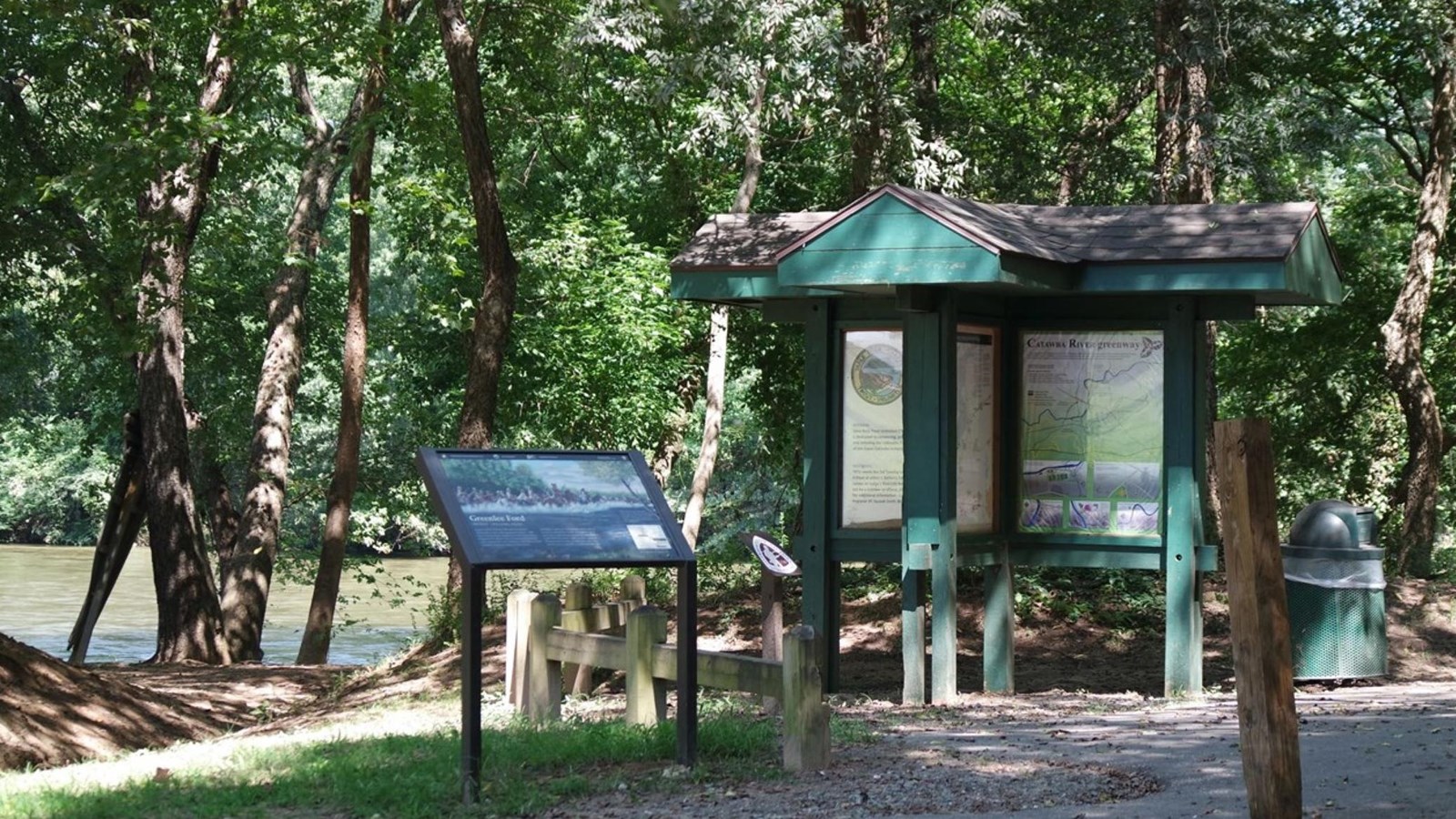 A large billboad and a park wayside are along a trail with trees and a river behind them. 