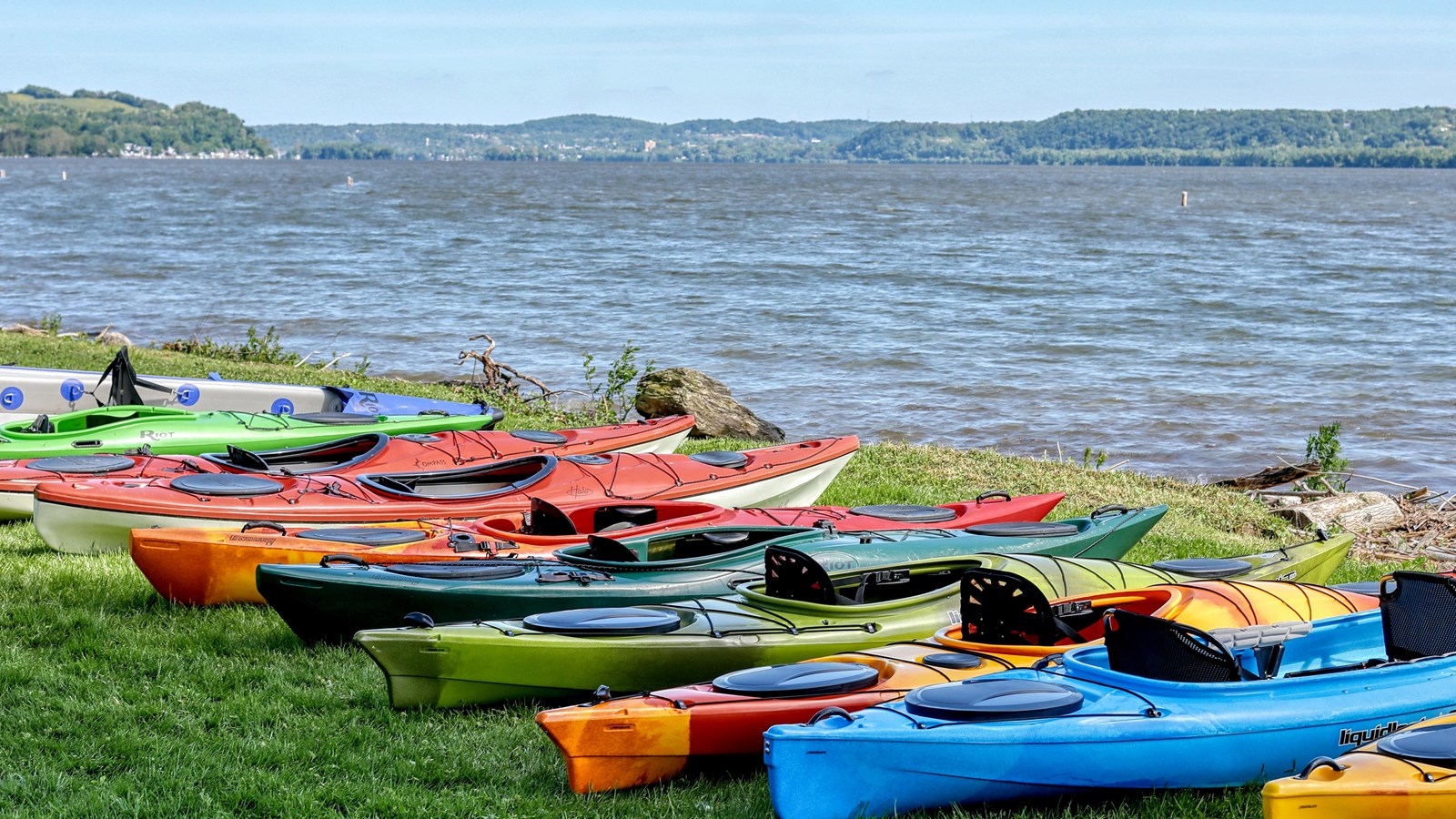 A variety of colorful kayaks along the grassy shore by a lake, with tree-covered hills in the back.