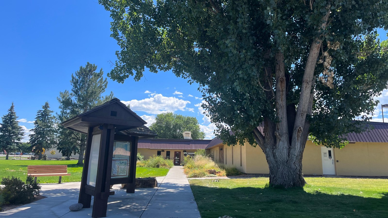 A brown exhibit sign next to a sidewalk leading into a yellow, one-story buidling.
