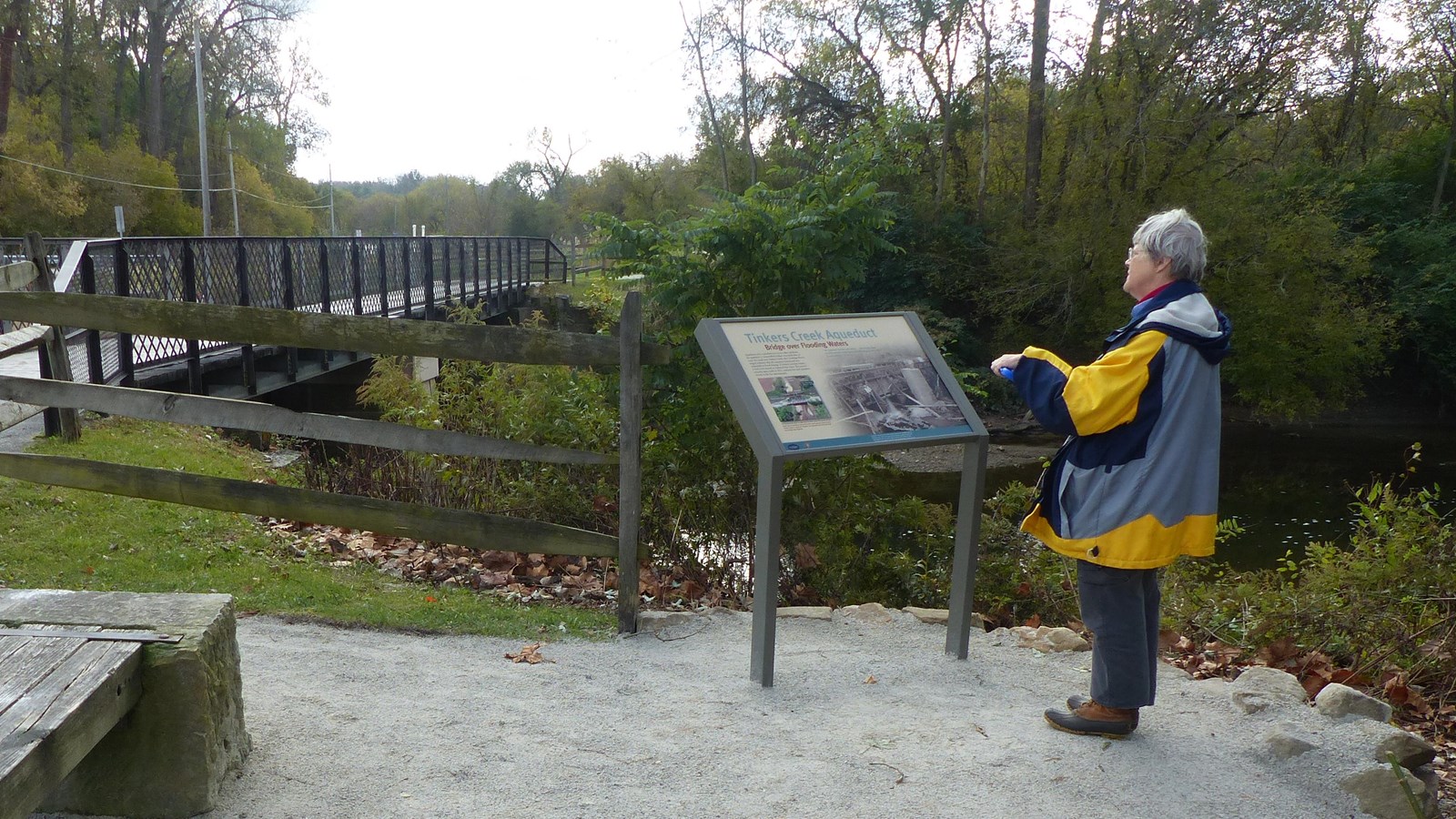 A woman in a raincoat reads a graphic panel by a stream. 