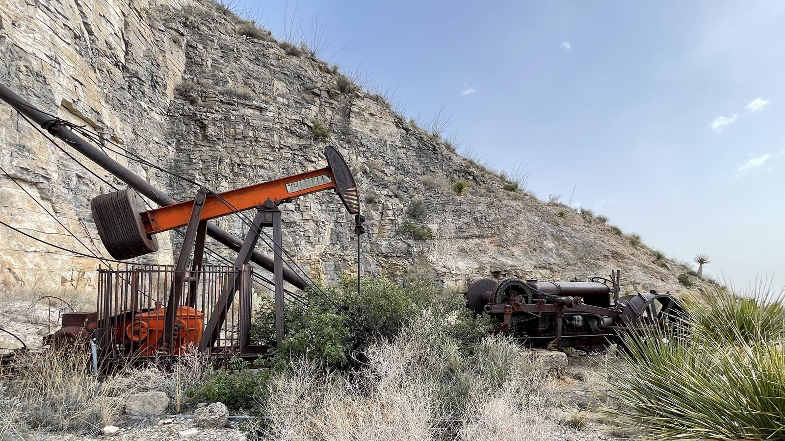 An abandoned oil pump jack stands in a flat space on a rugged desert mountainside.