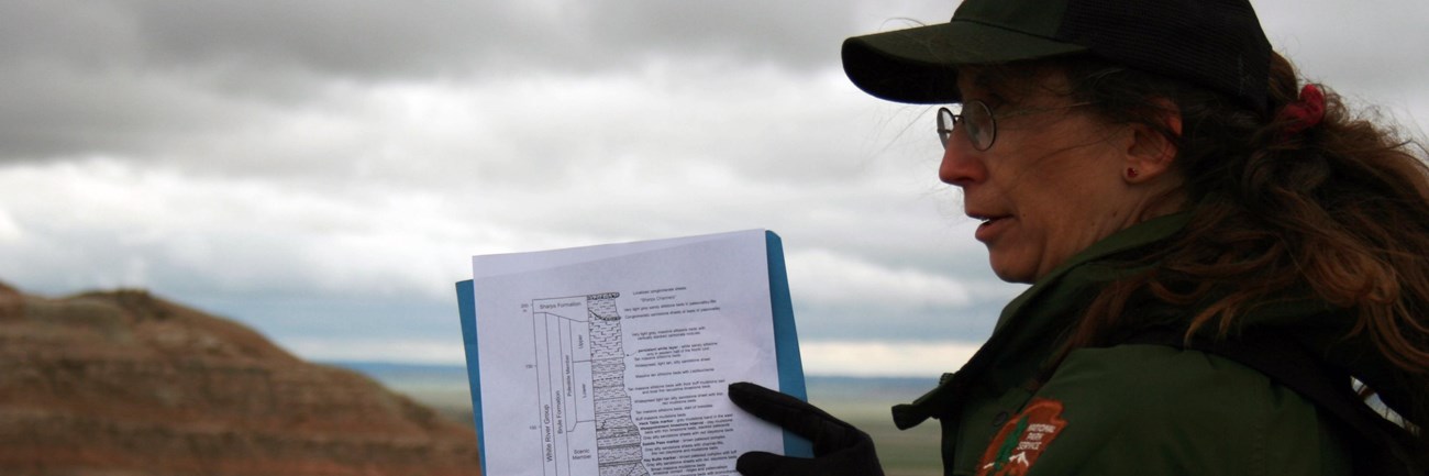 a ranger points at a geologic column on a piece of paper in front of a badlands butte.