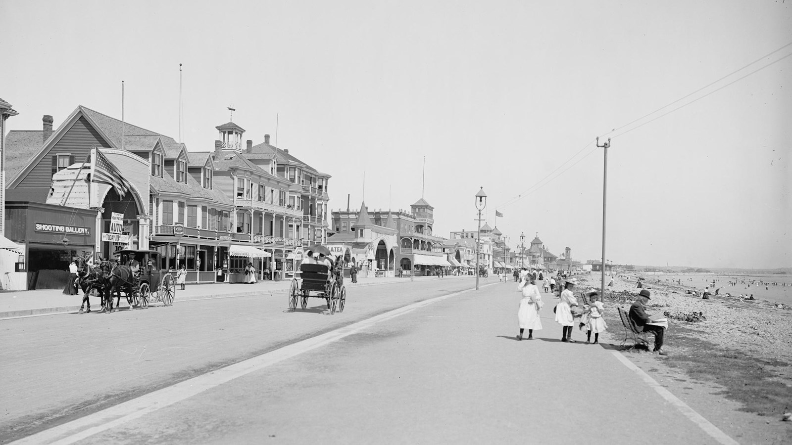 Ealry 20th century photo of people walking along a boardwalk with beach on the right. 