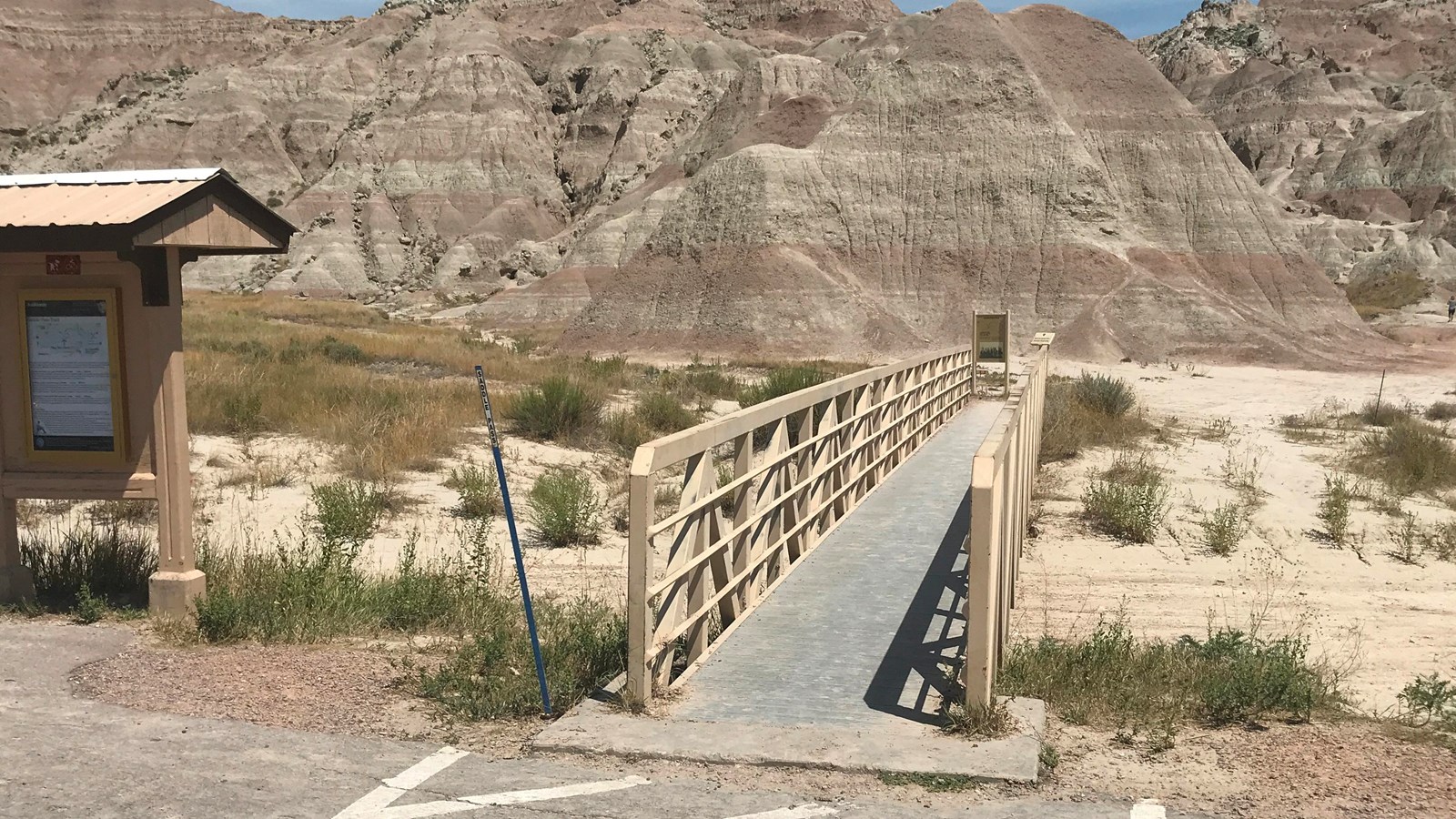 Information board sits alongside boardwalk which extends into badlands formations under blue sky.