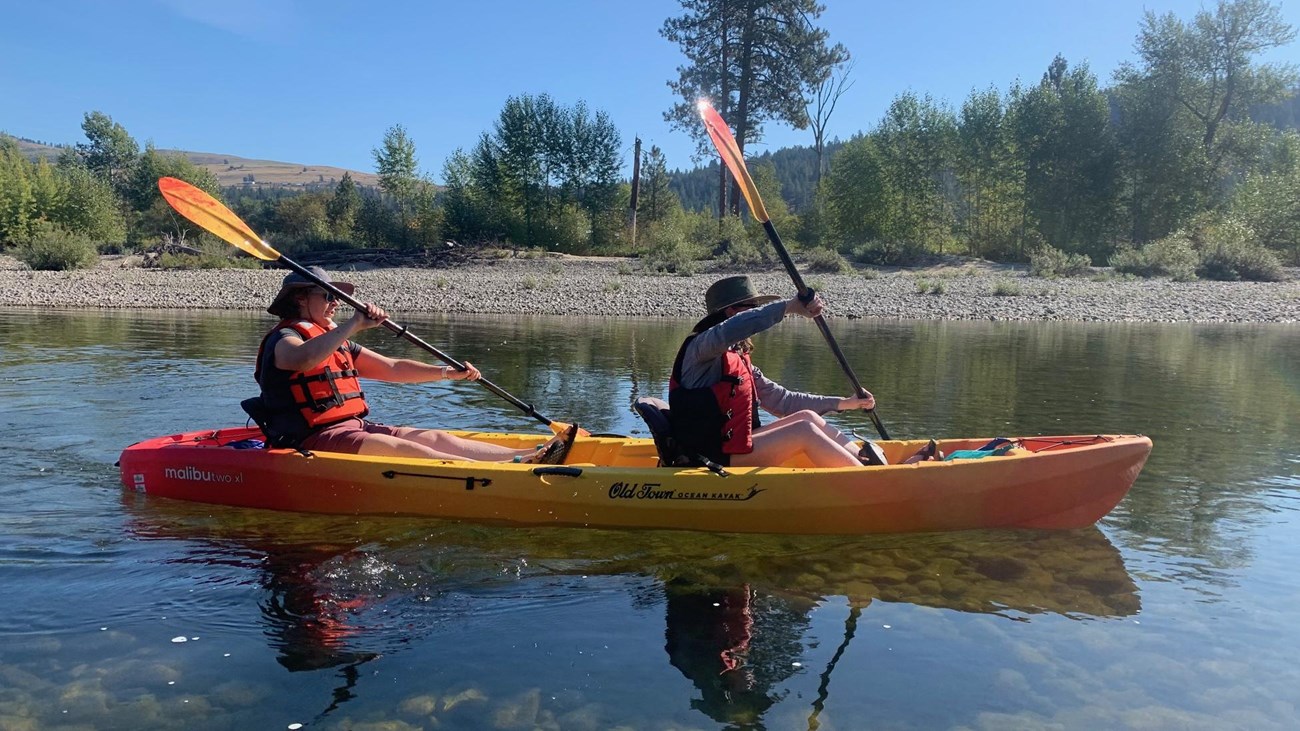 An orange kayak holds two paddlers out on clear shallow water.