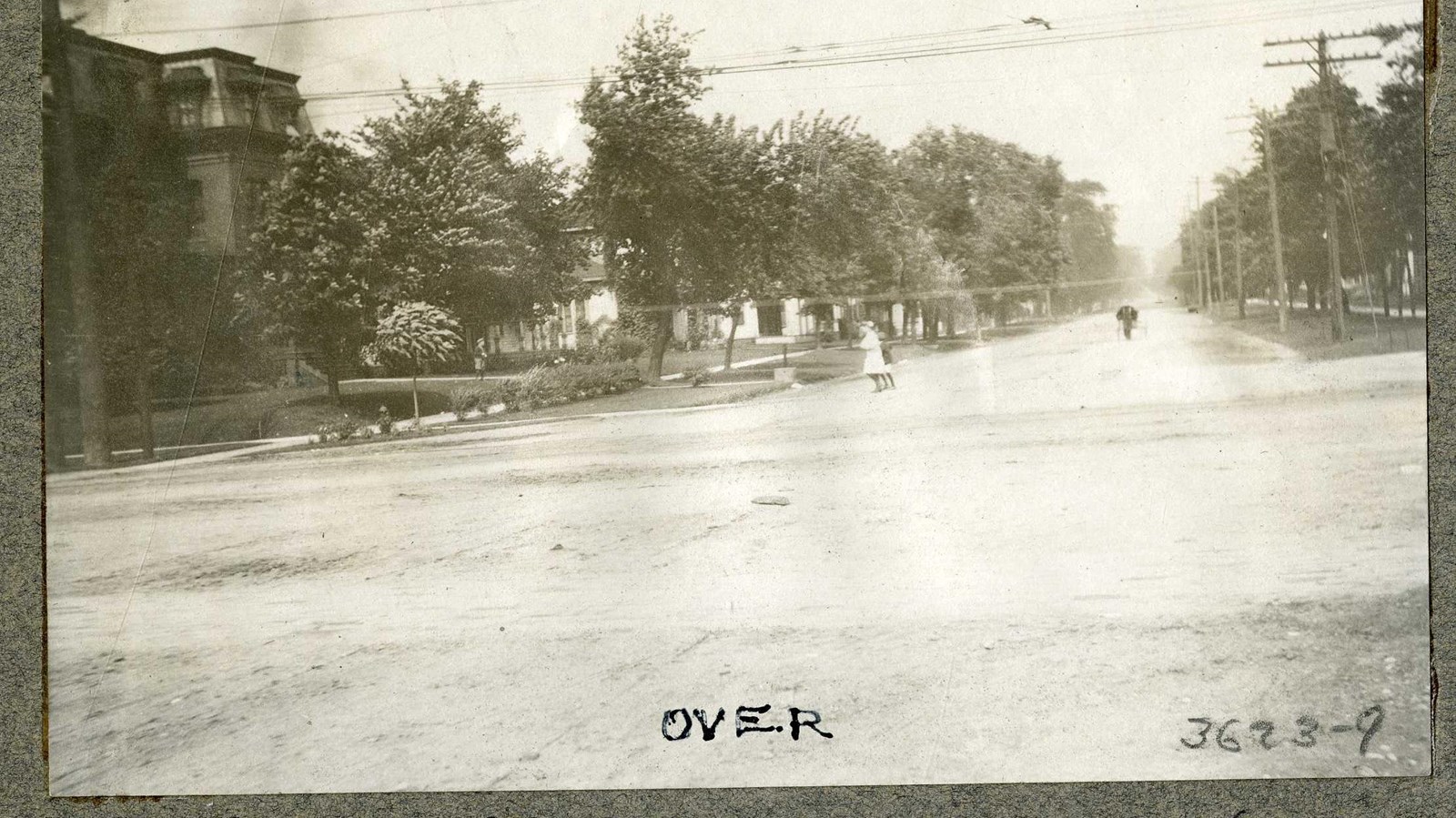 Black and white of flat road lined with trees, grass, and homes, people along the road 