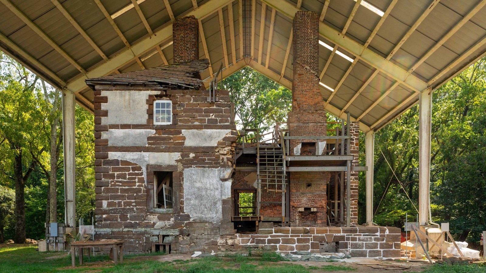 Partially reconstructed historical building with a chimney under a metal roof structure amidst trees