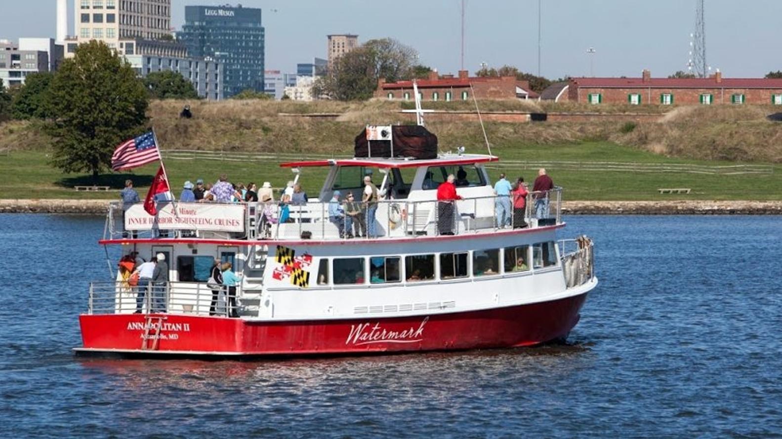 A red and white sightseeing boat with passengers near Fort McHenry and a city skyline in the backgro