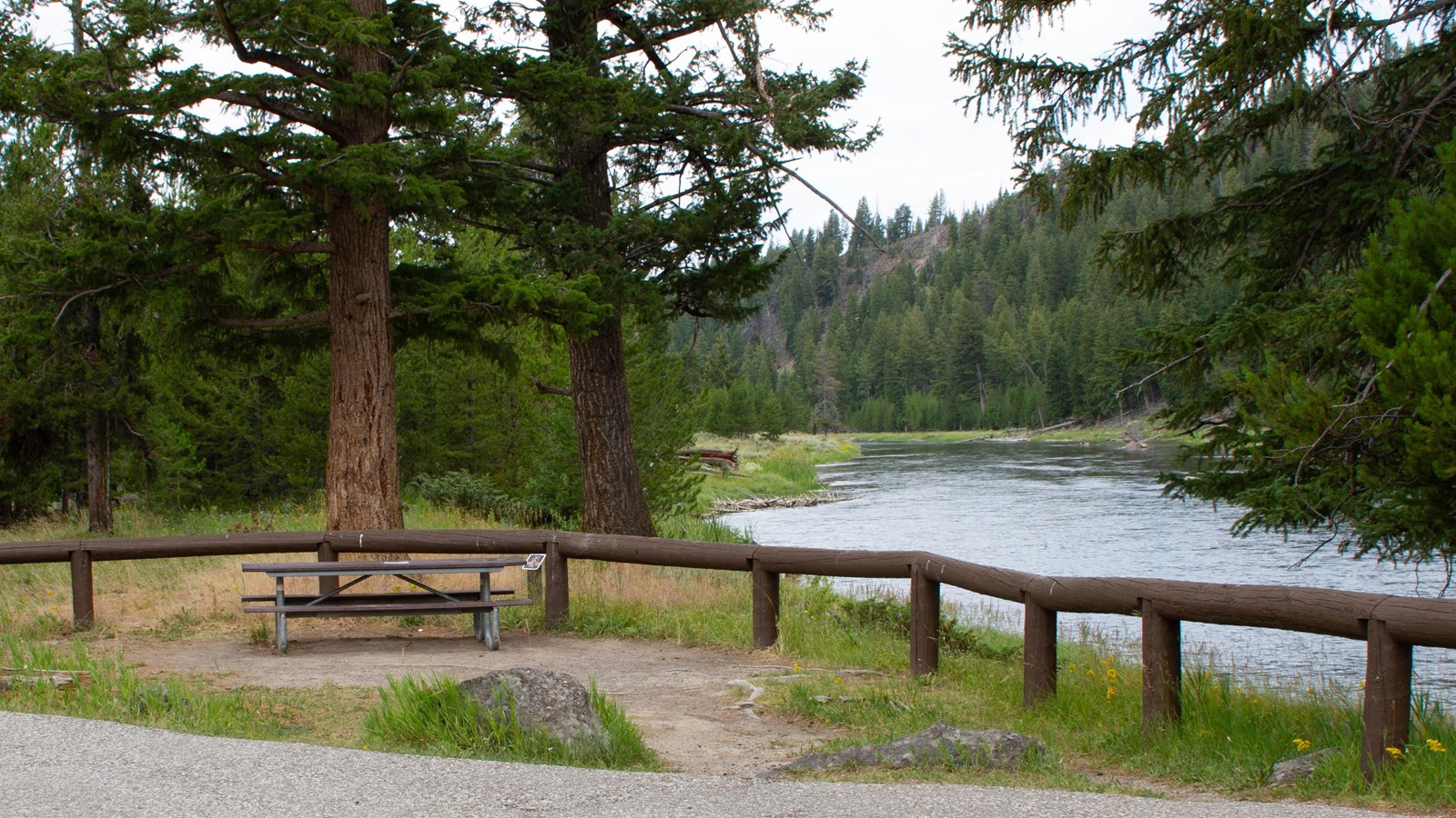 A picnic table in front of a wooden fence near the shore of a river.