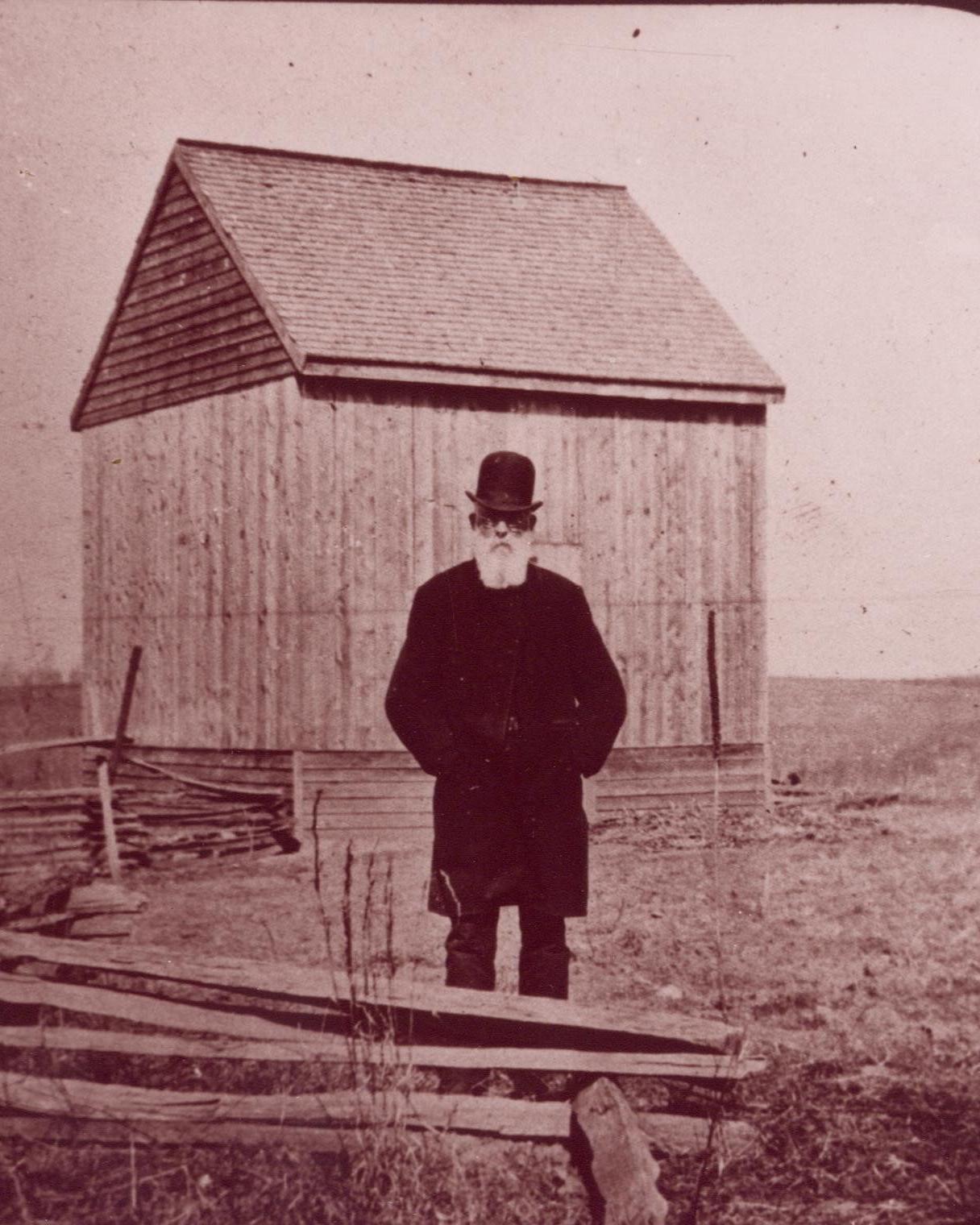 Older man with a white beard and hat with a wooden barn in the background. 