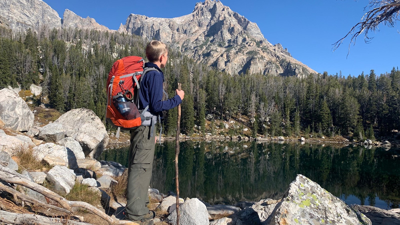 Backpacker standing at the edge of an alpine lake with mountains and trees surrounding
