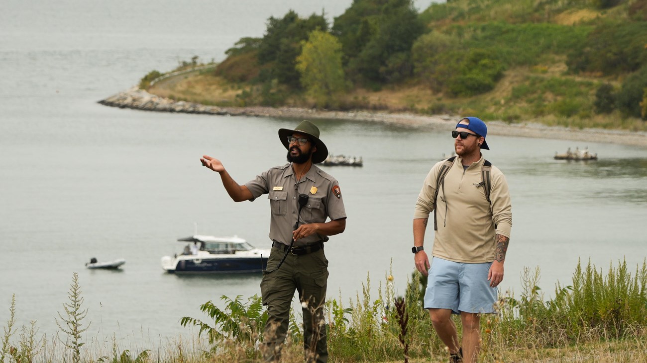 A uniformed ranger walks with a guest on a grassy path on and island, boats in the water behind them