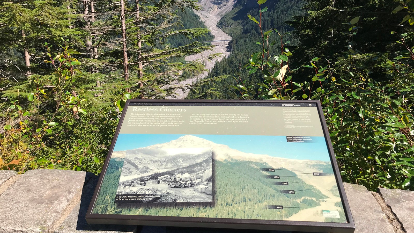 A exhibit panel overlooking a forested valley with a wide rocky river bed. 