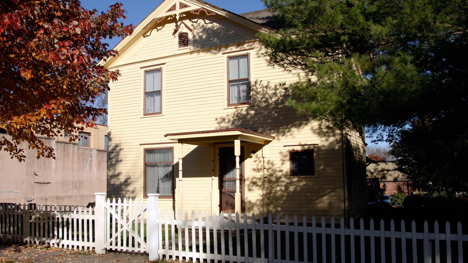 A yellow house with brown trim and a covered front porch is partially shaded by trees.