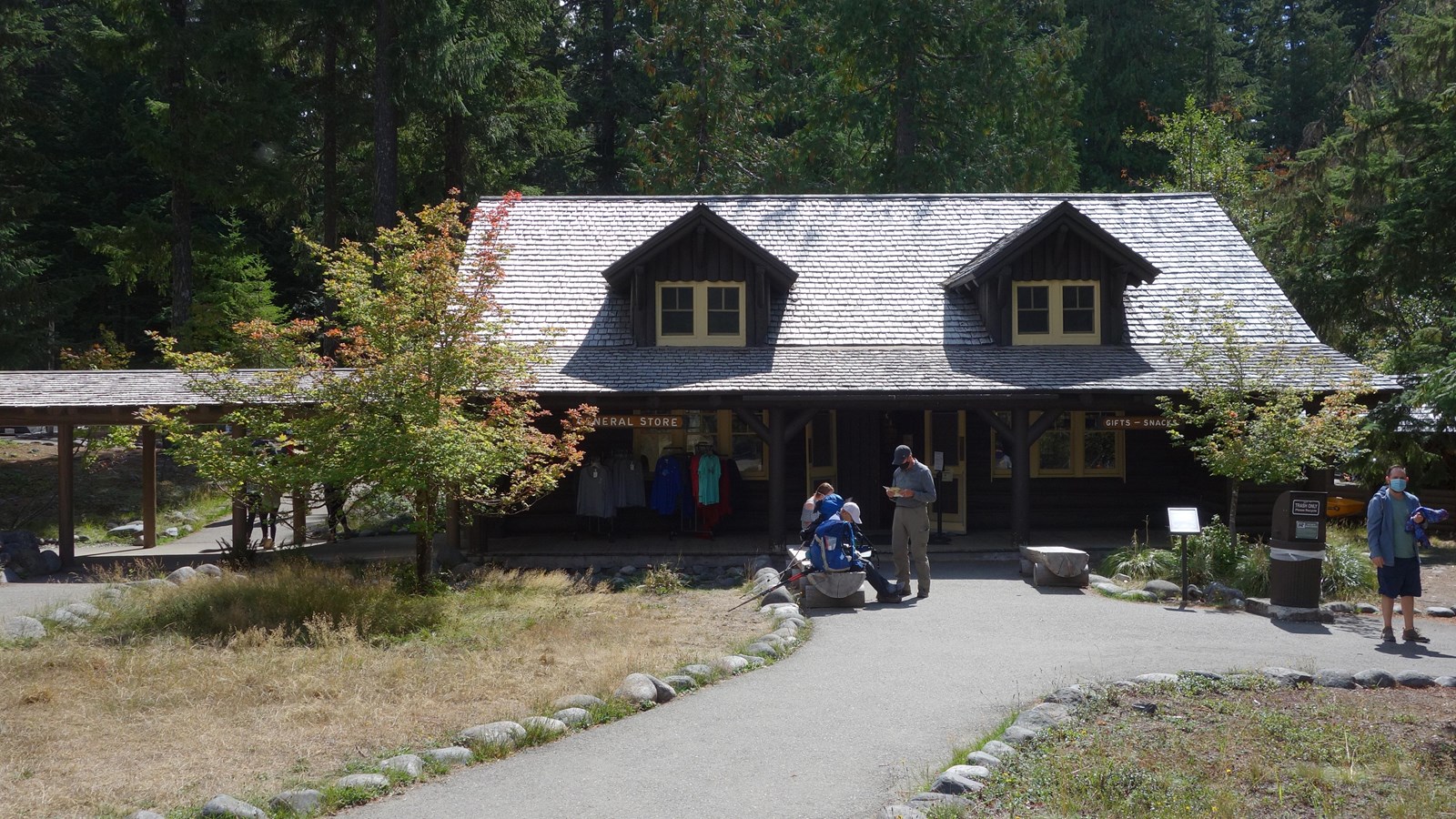 A small wayside panel next to a paved path in front of a wood building with people on the path.