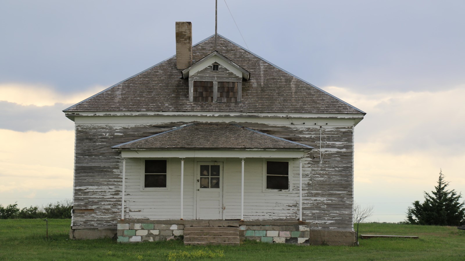 White frame schoolhouse with a front porch on cement brick foundation in the middle of a field.