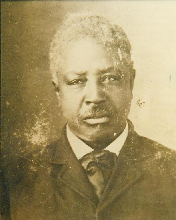 Sepia photo of a man with short white hair and mustache, wearing a black suit and black tie.