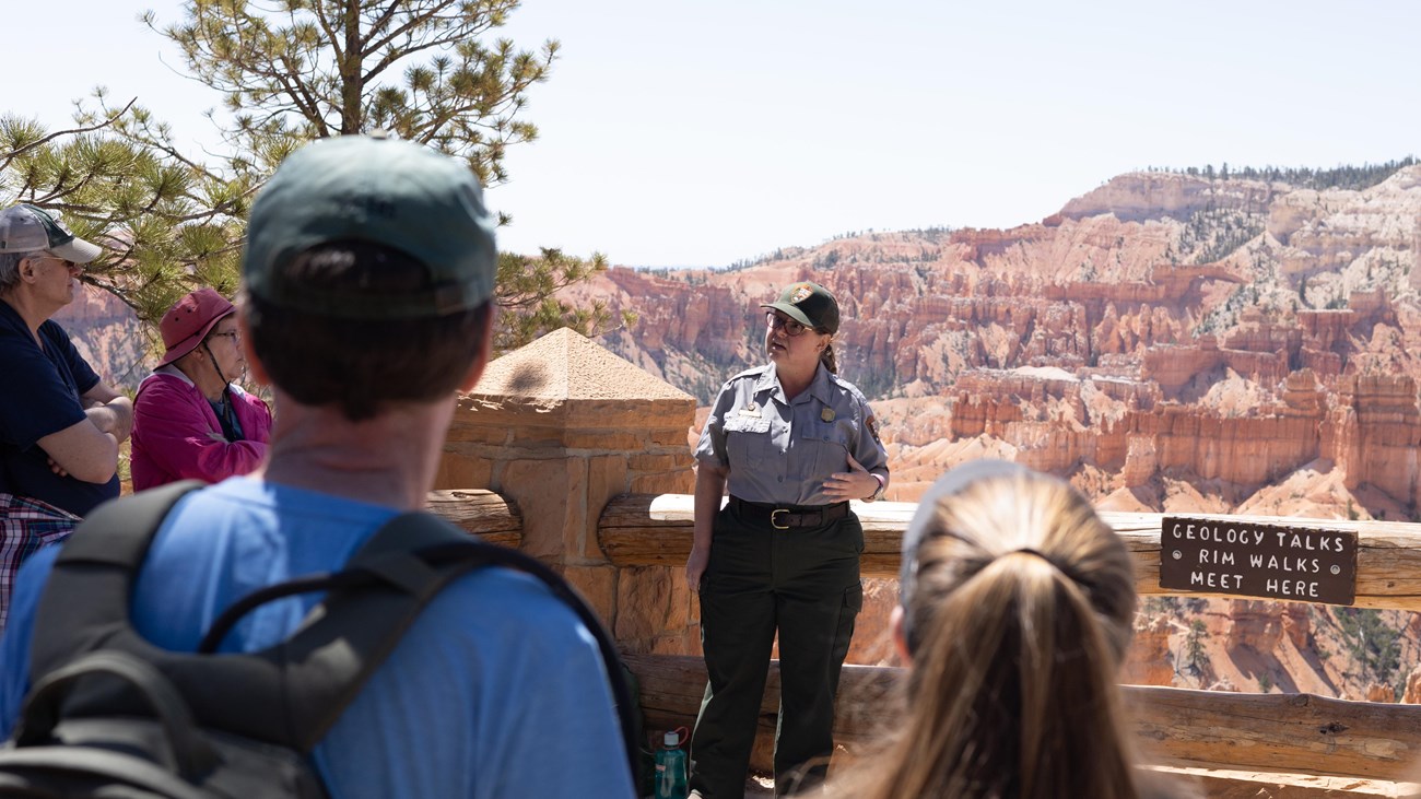 A ranger in uniform stands in front of a crowd with a background of red rock formations