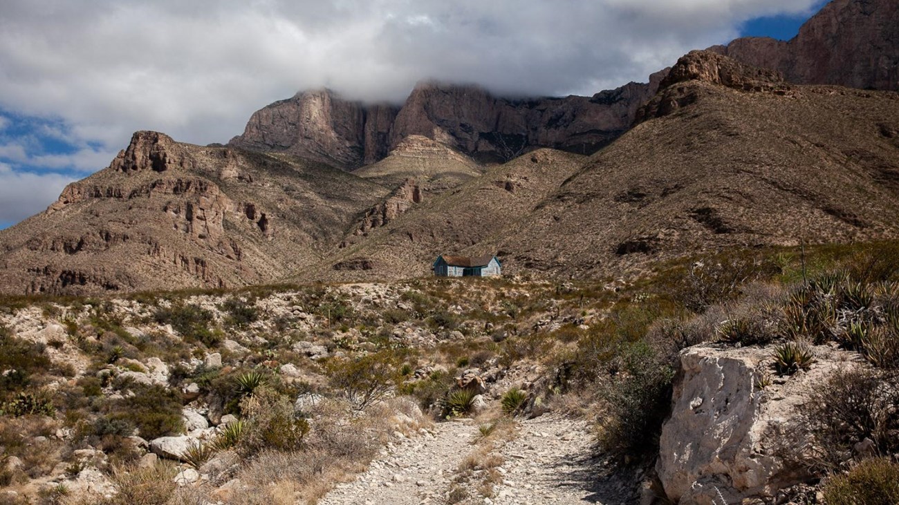 A primitive dirt road leads to a small blue ranch house dwarfed by desert mountains. 