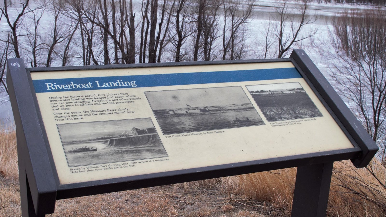 Exhibit stands on rivers edge, trees, water and winter river ice behind sign.
