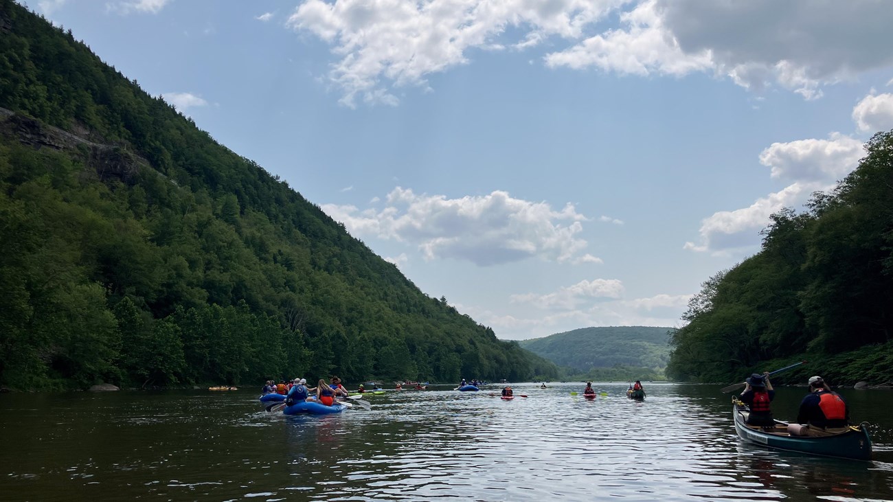Group of rafts and canoes paddle on river in between forested two mountains/hills.