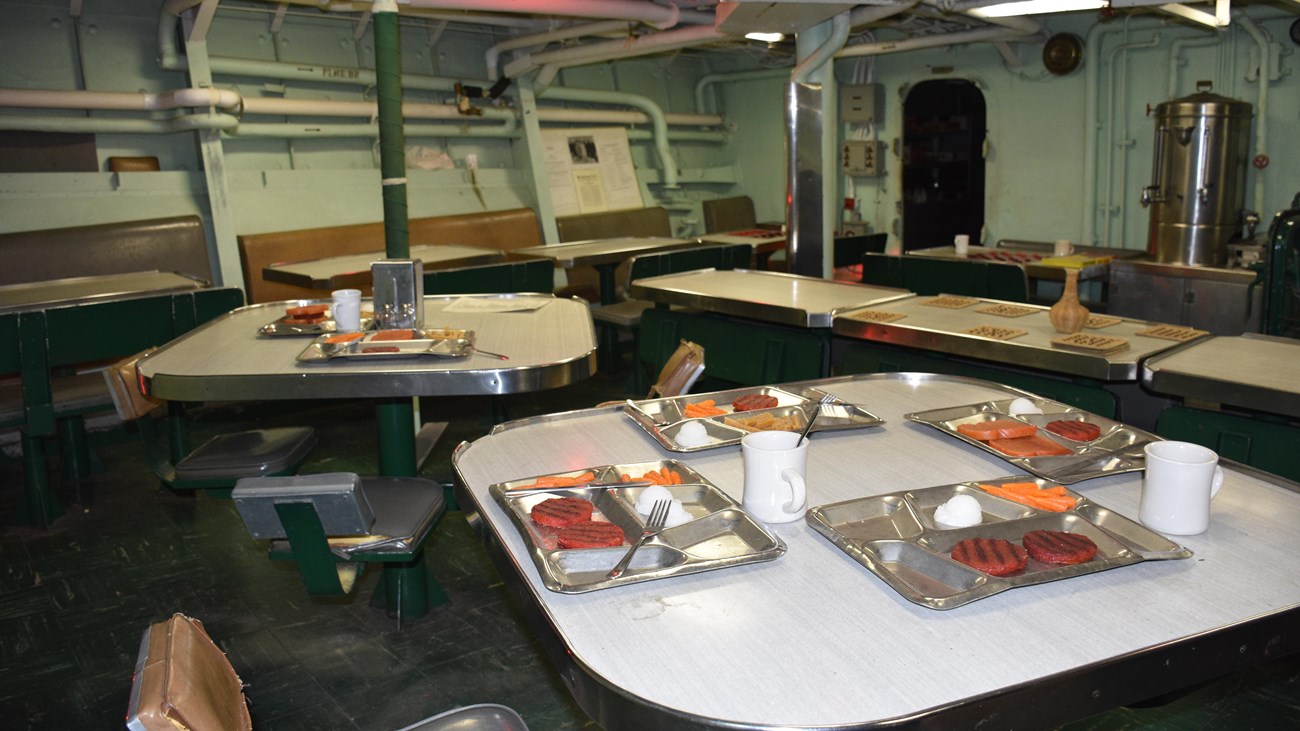 The mess of the USS CASSIN YOUNG which has white square tables with gray food trays on top. 