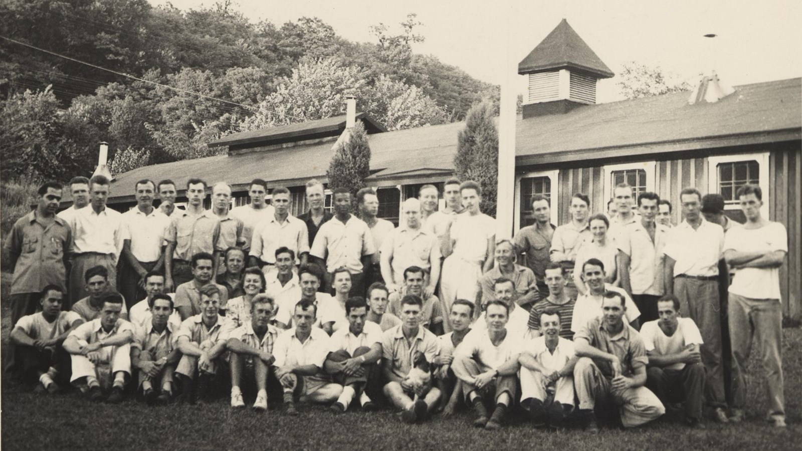 Group photo of about fifty people, mostly men, standing outside in front of a building and trees.