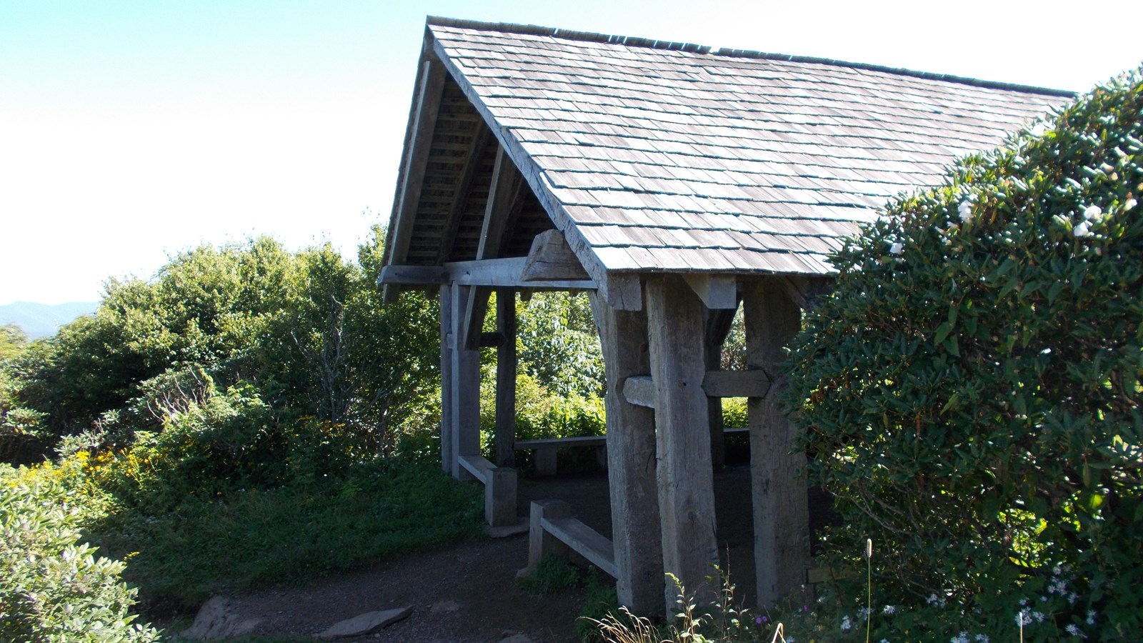 Large wooden trail shelter with shake roof and open sides surrounded by shrub vegetation