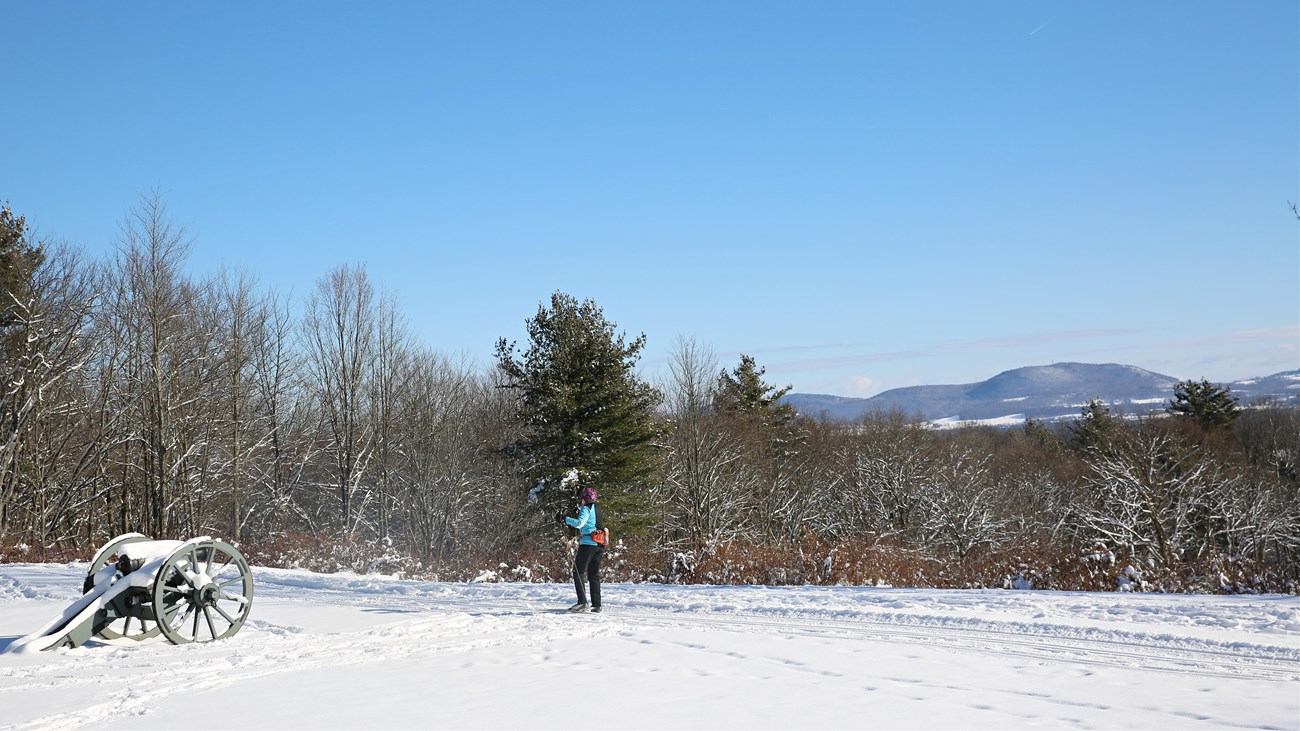 A cross-country skier traverses down a groomed path on a hilltop and past a cannon covered in snow.