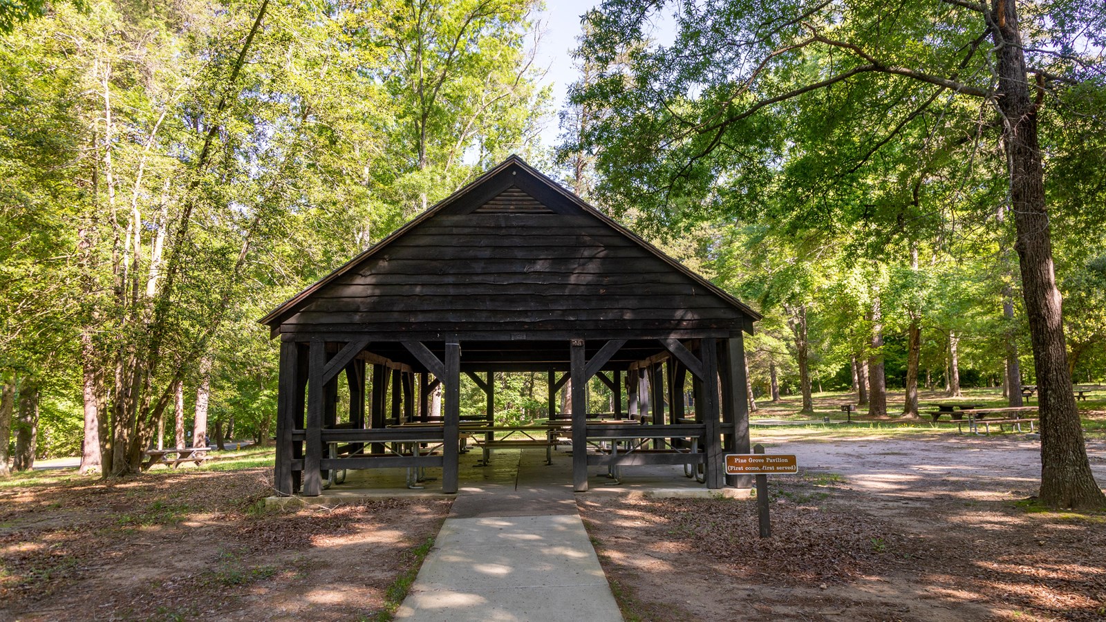A wooden pavilion with wooden picnic tables sits among the woods. A sidewalk leads up to it.