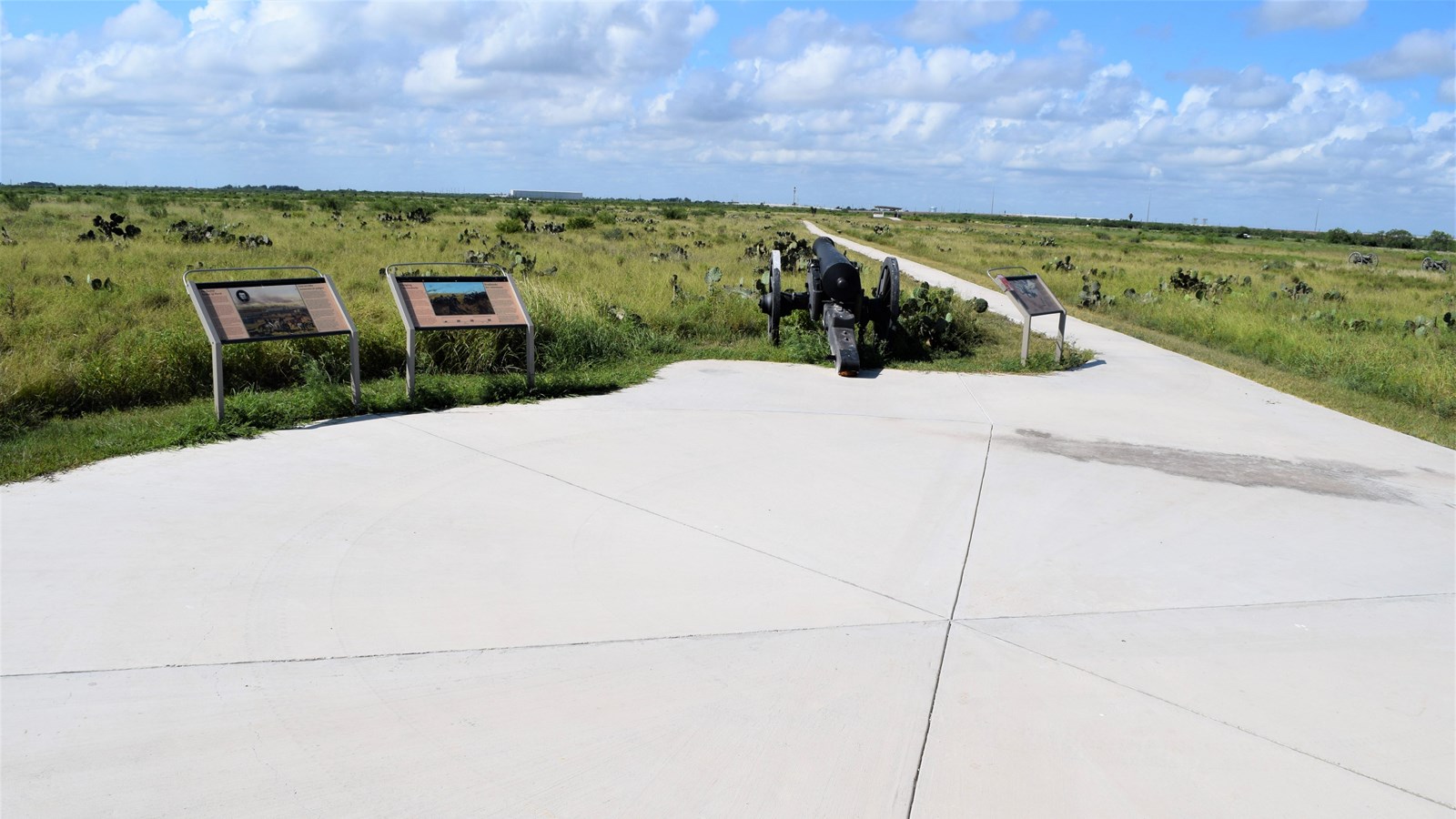 A paved concrete walking trail with replica cannon and waysides cutting through the coastal prairie.