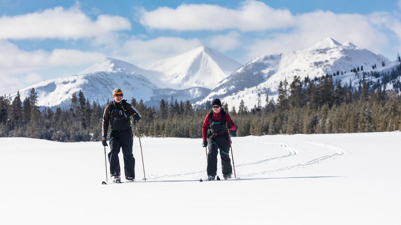 Two skiers travel across a flat meadow with mountains in the distance.