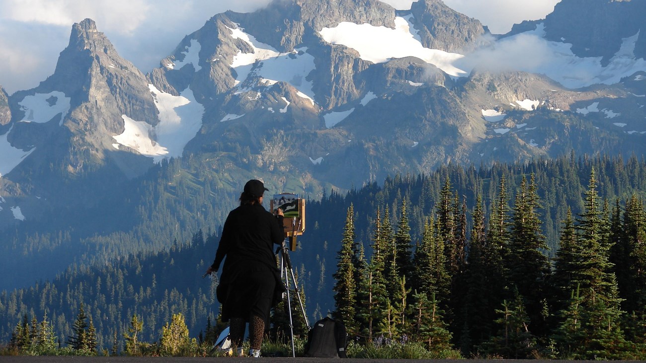 A female figure viewed from behind stands at an easel painting a picture of the tall mountains in fr