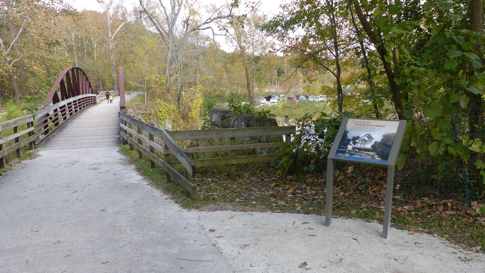 A trail leads by a graphic panel and stone wall, and crosses a pedestrian bridge to a parking lot.