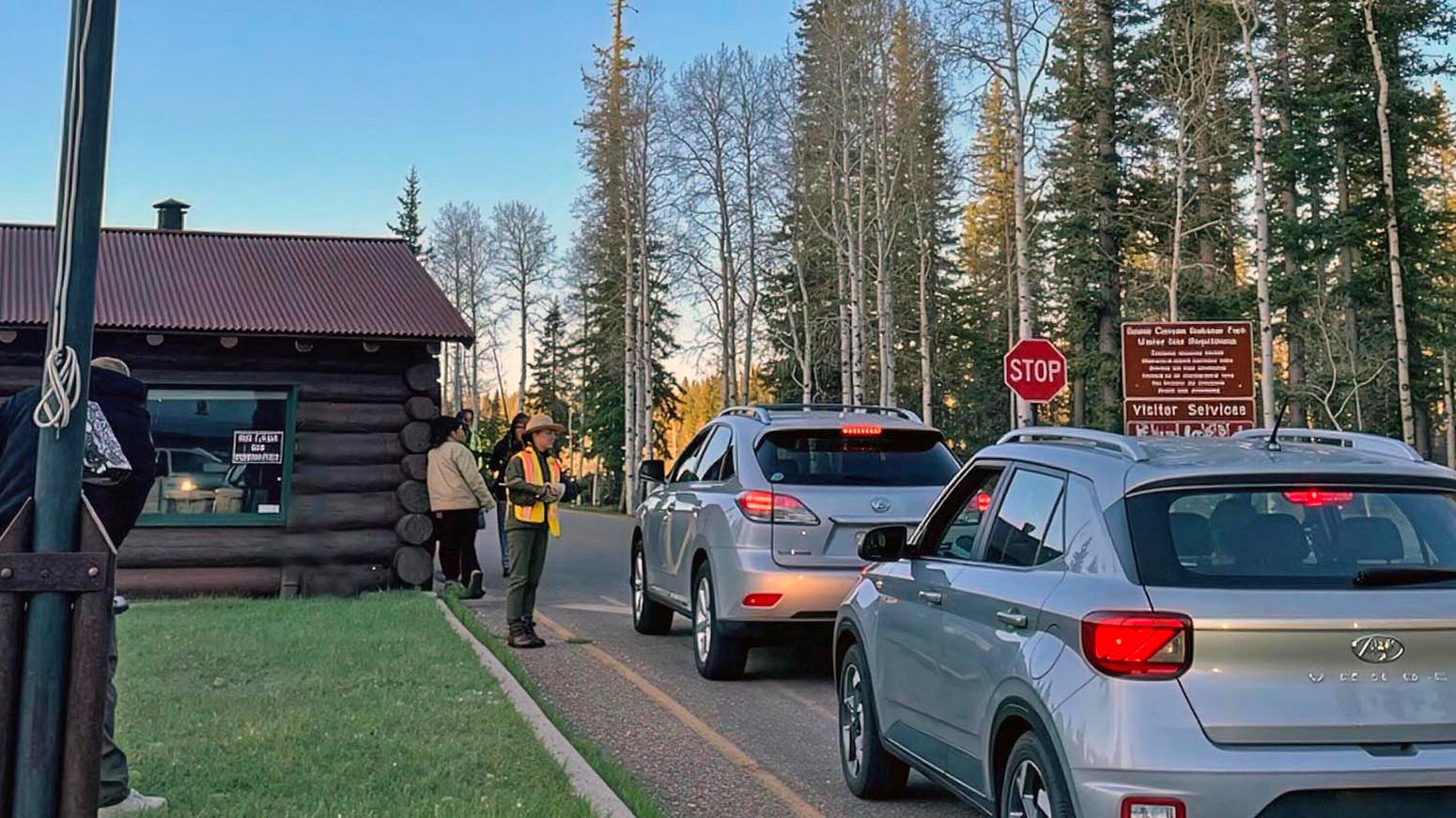 Two cars in line at a rustic log entrance station. A park ranger is greeting them and providing maps